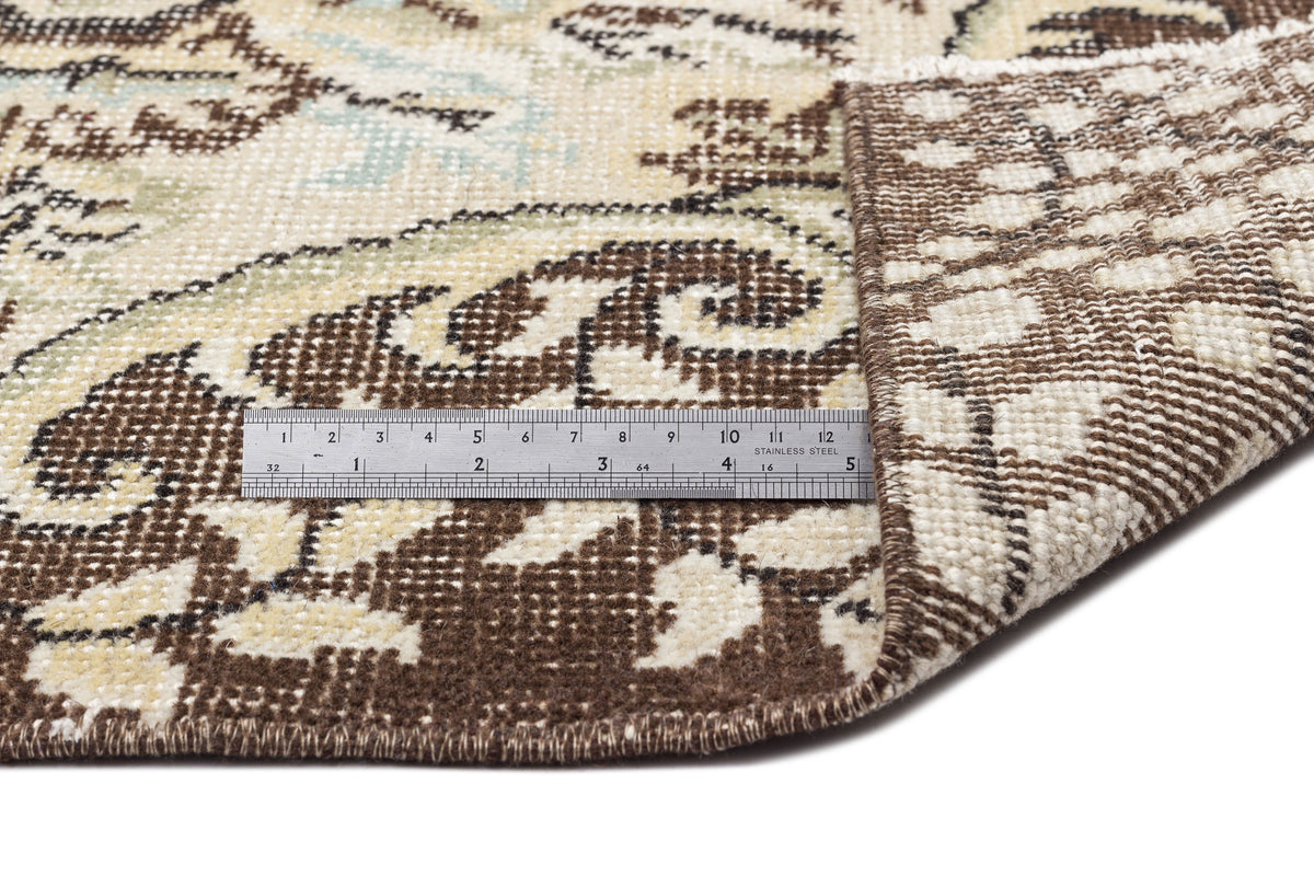 Brown, cream, and sage green floral patterned rug with a folded corner showing the weave and a metal ruler for scale.
