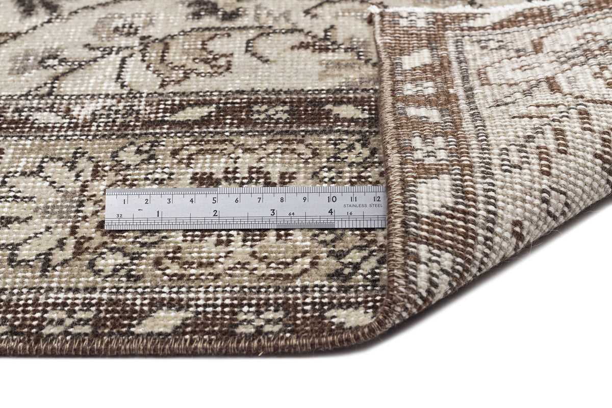 A close-up shows the corner of the Sigima - Vintage Floral Rug (5'4 x 9'1) flipped over to reveal its back, with a metal ruler displaying cm and inch markings for scale. The rug features brown, beige, and cream vintage-inspired designs.