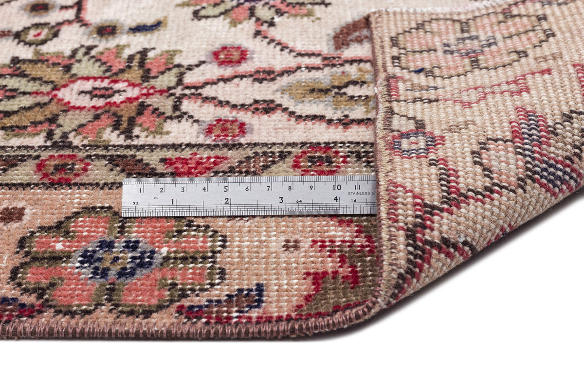 Beige, pink, and green floral patterned rug with a corner folded back, showing the woven backing and a metal ruler.