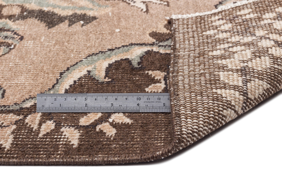 Brown and beige floral patterned rug with a folded corner showing the woven backing and a metal ruler for scale.