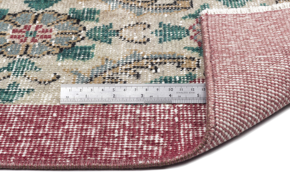 Beige, green, and red floral medallion rug with a folded corner and a metal ruler for scale on a white background.