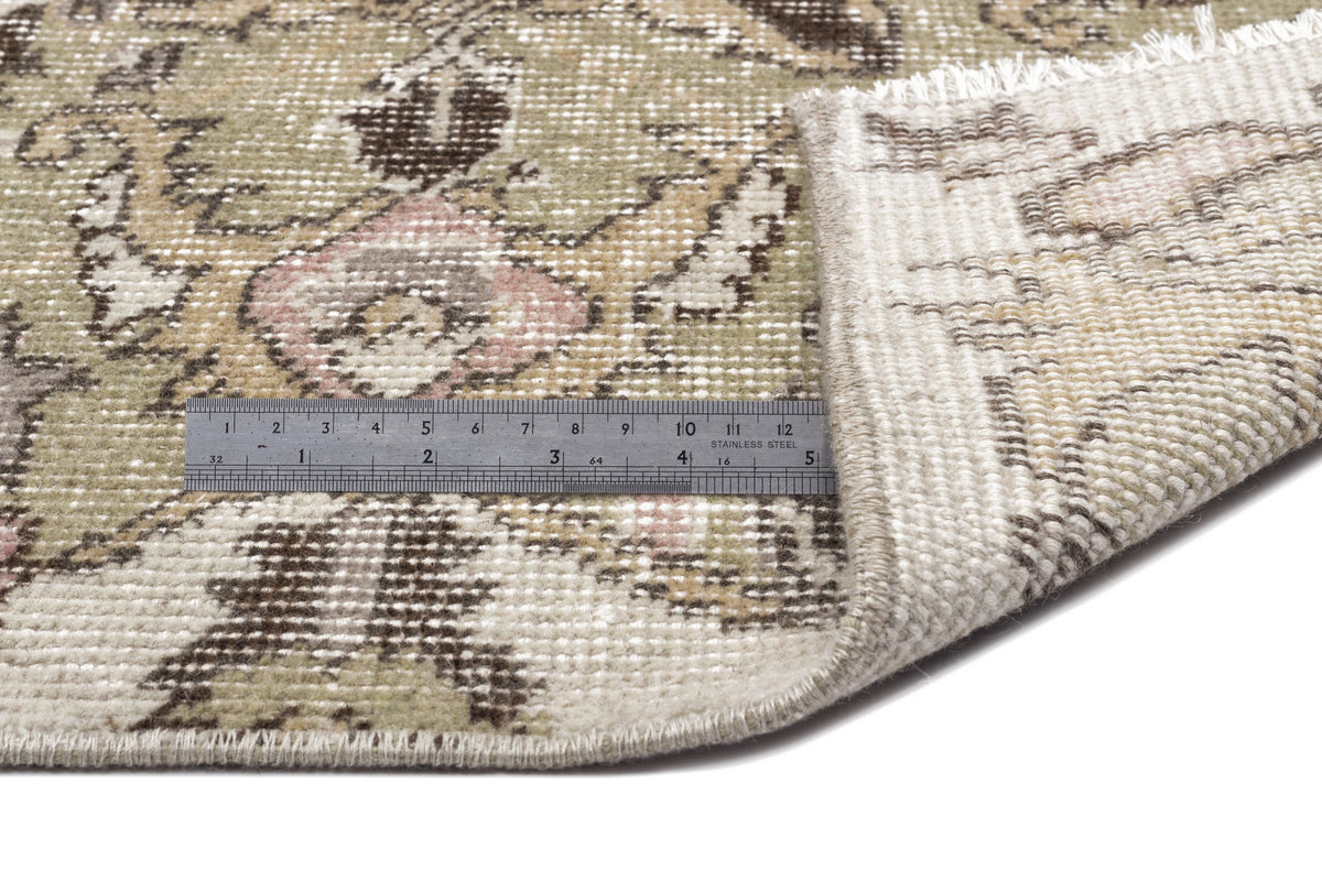 A close-up of the Kaugila - Vintage Medallion Rug (5'9 x 9'0) corner flipped to reveal its underside, with a gray ruler for scale. The rug features floral patterns in muted beige, brown, and green tones.