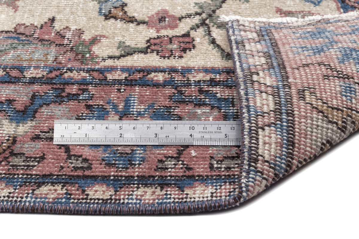 Beige, blue, and pink floral patterned rug with a folded corner revealing the woven backing and a metal ruler for scale.