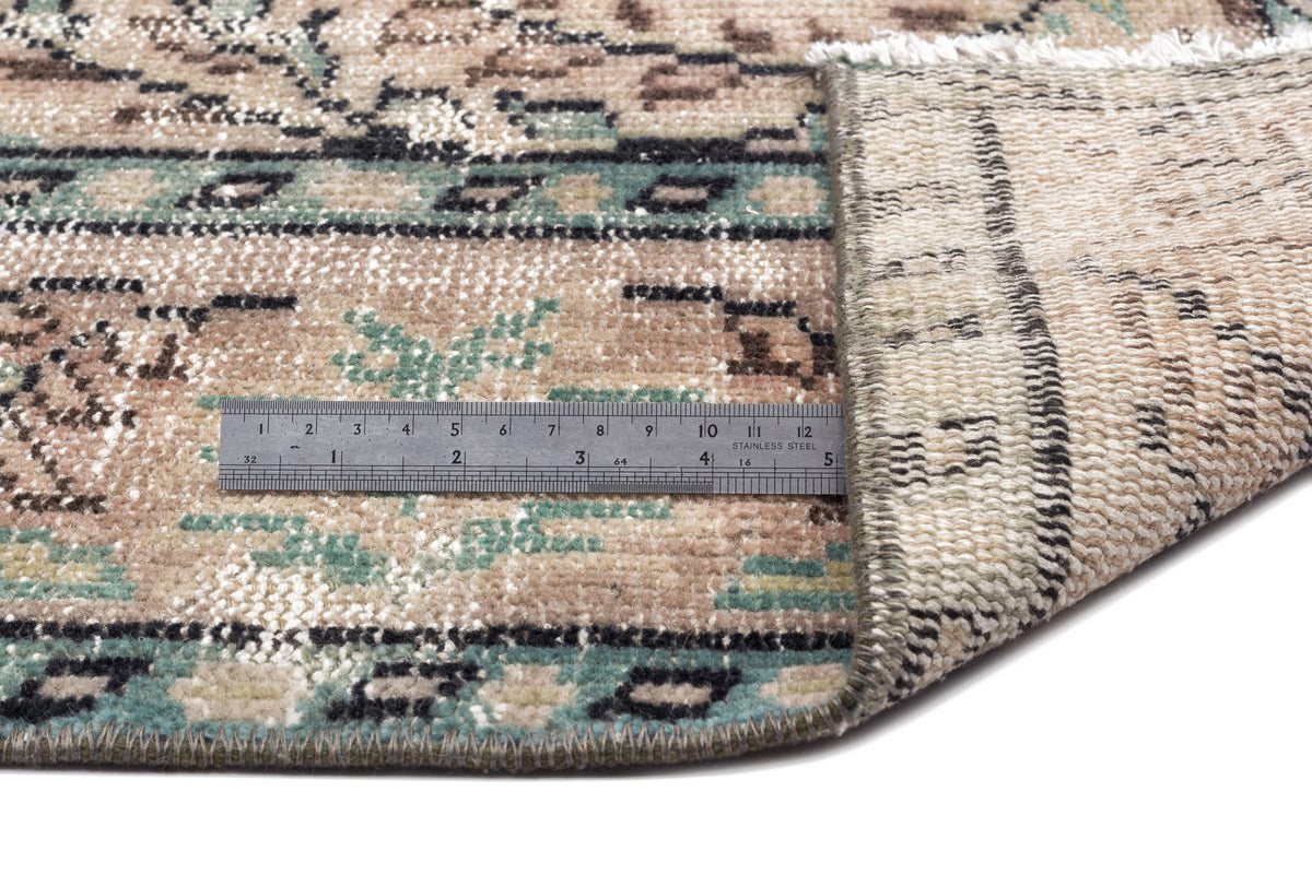 Beige, sage green, and black distressed patterned rug with a folded corner and a metal ruler for scale.