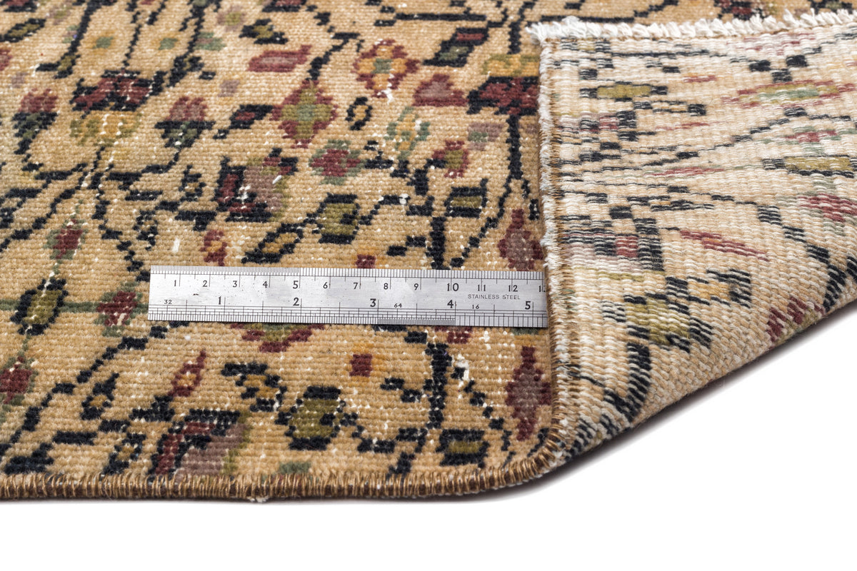 Tan rug with a multi-colored floral pattern and a folded corner showing the backing, measured by a metal ruler.