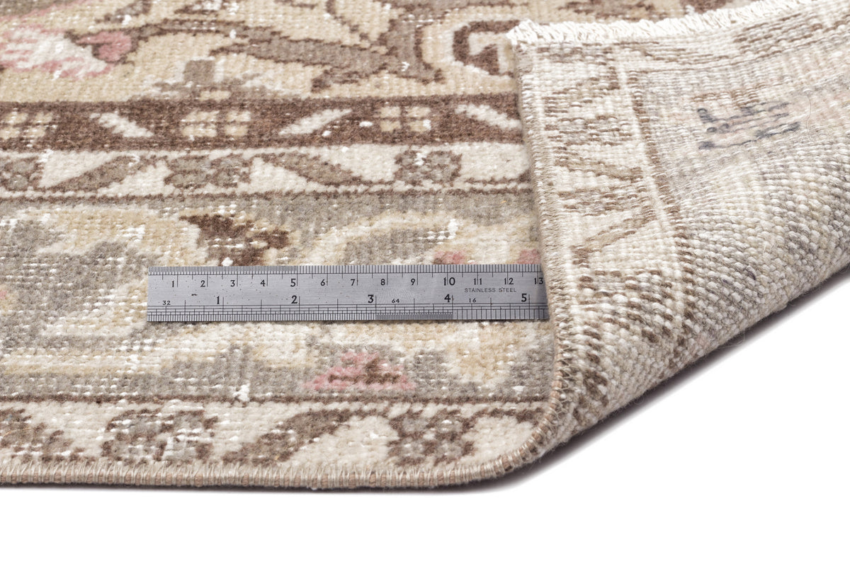 Beige and brown vintage-style rug with floral motifs and a folded corner, shown with a metal ruler for scale.
