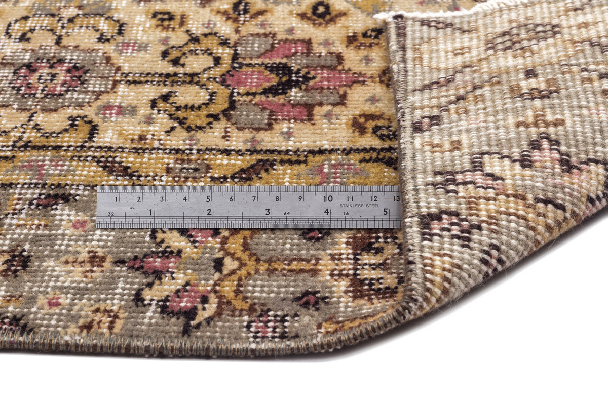 Distressed floral medallion rug in gold, grey, and pink with a metal ruler showing pile height on a white background.