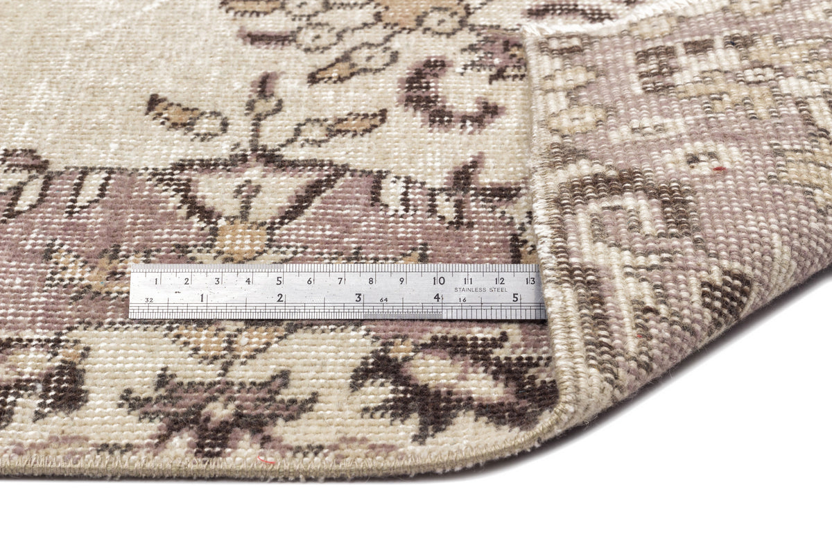Distressed beige and mauve vintage-style rug with a floral medallion pattern, shown with a metal ruler for scale.