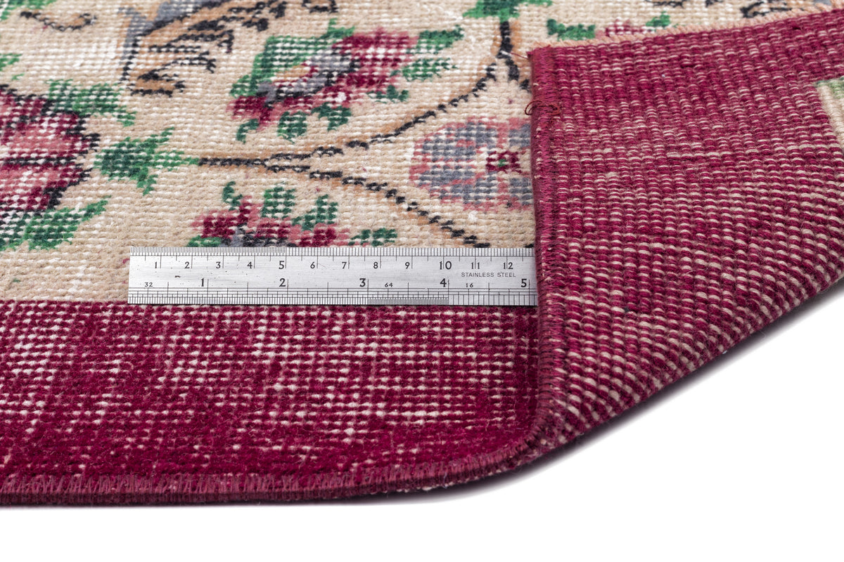 Beige and burgundy floral rug with a folded corner showing the woven backing and a metal ruler for scale.