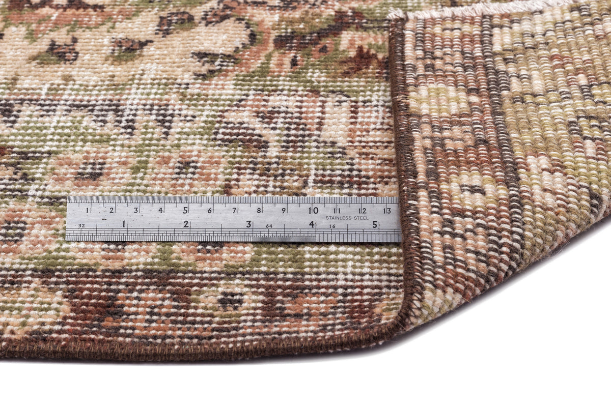 Close-up of a brown, green, and beige floral patterned rug with a metal ruler showing scale, isolated on white.