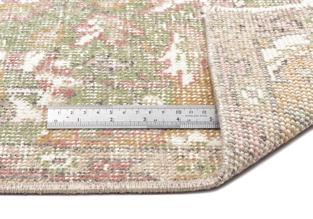 A close-up of the Bofaya - Vintage Medallion Rug (6'4 x 10'2) with a metal ruler in centimeters on top, showing its beige, green, pink, and white pattern. One corner of the rug is folded over.