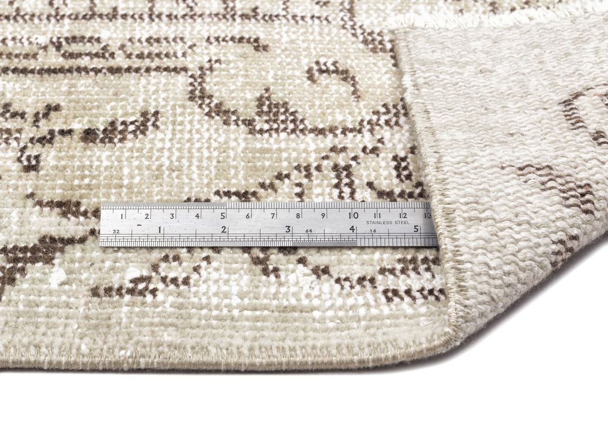 Beige and brown vintage-style rug with a floral pattern, showing a corner fold and a metal ruler for scale.