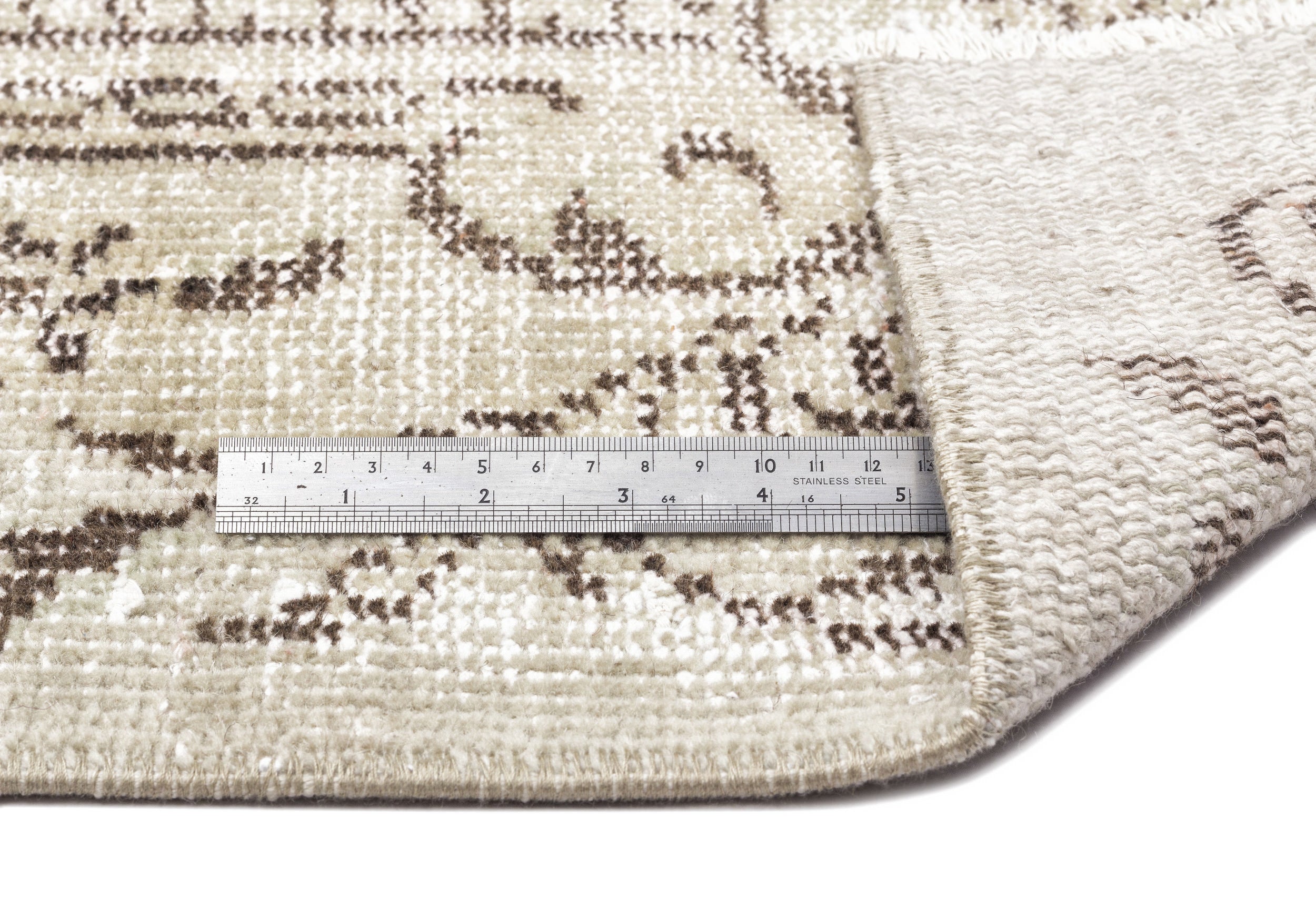 Beige and brown vintage-style rug with a floral pattern, showing a corner fold and a metal ruler for scale.