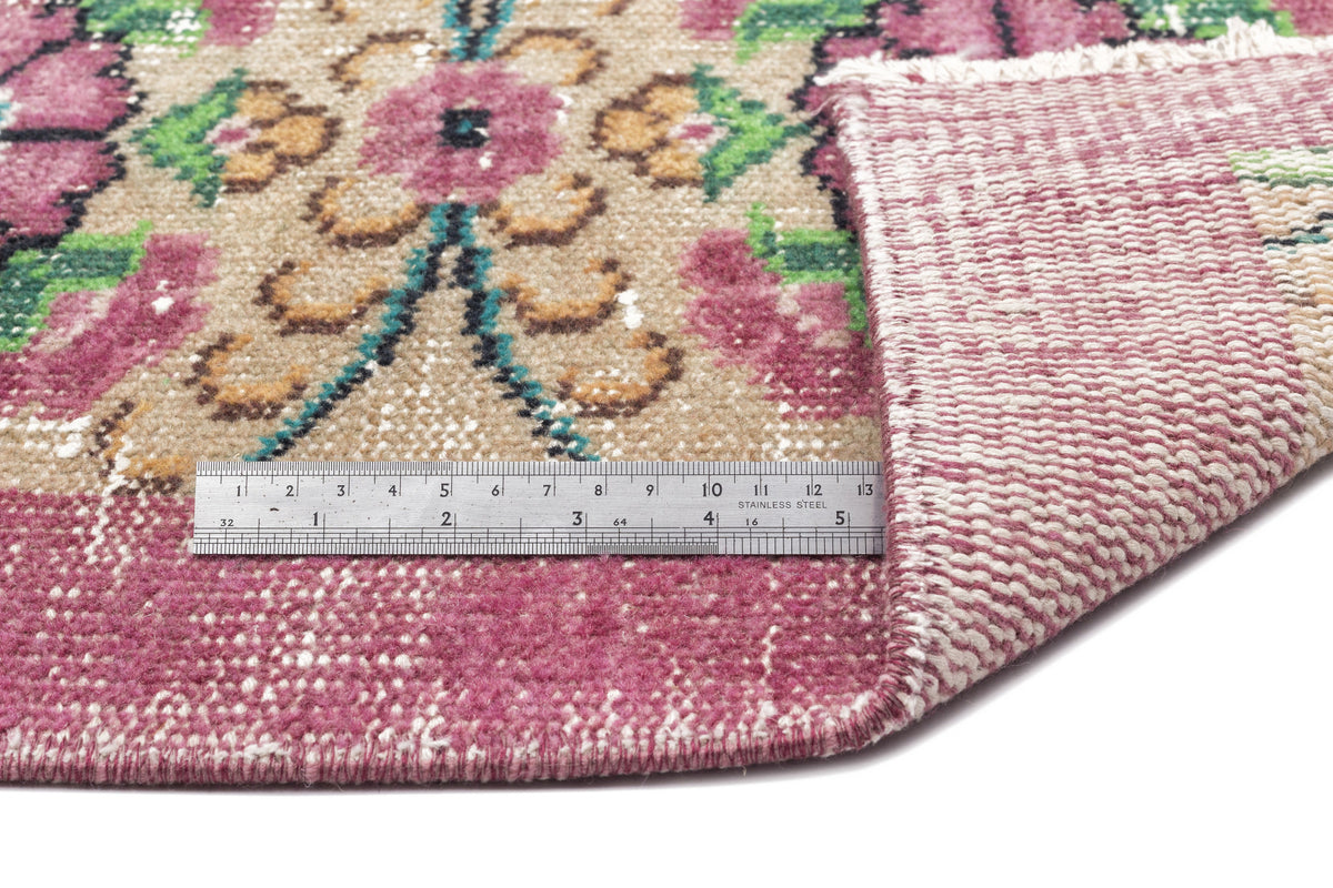 Distressed floral rug in magenta, tan, and green with a folded corner and a metal ruler for scale on a white background.