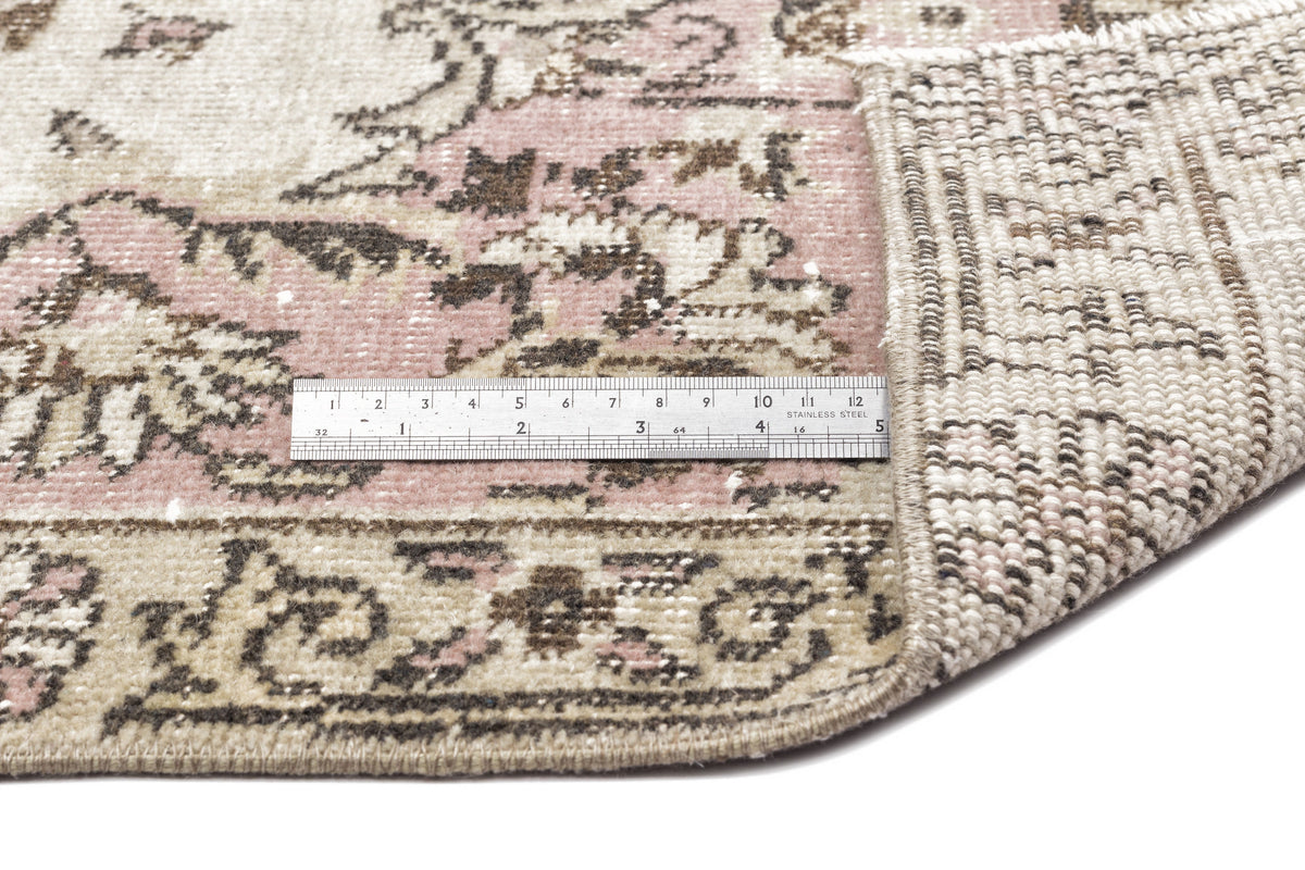 Distressed pink, beige, and brown vintage-style rug with a floral medallion pattern, showing the woven backing and a ruler.