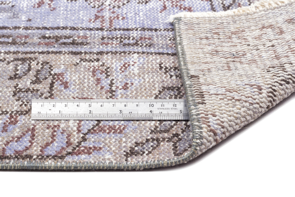 Distressed lavender and brown patterned rug with a folded corner and a metal ruler for scale on a white background.