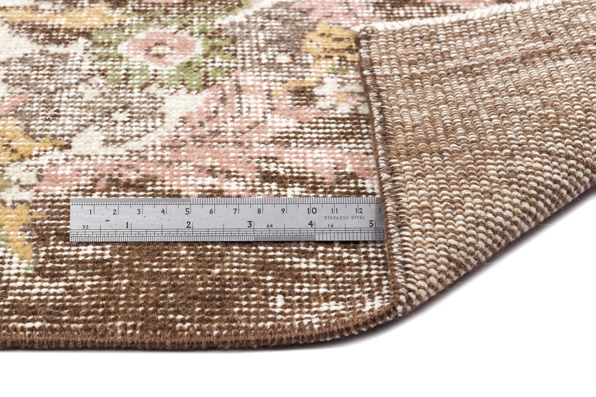 Brown, pink, and yellow floral patterned rug with a folded corner and a metal ruler for scale on a white background.