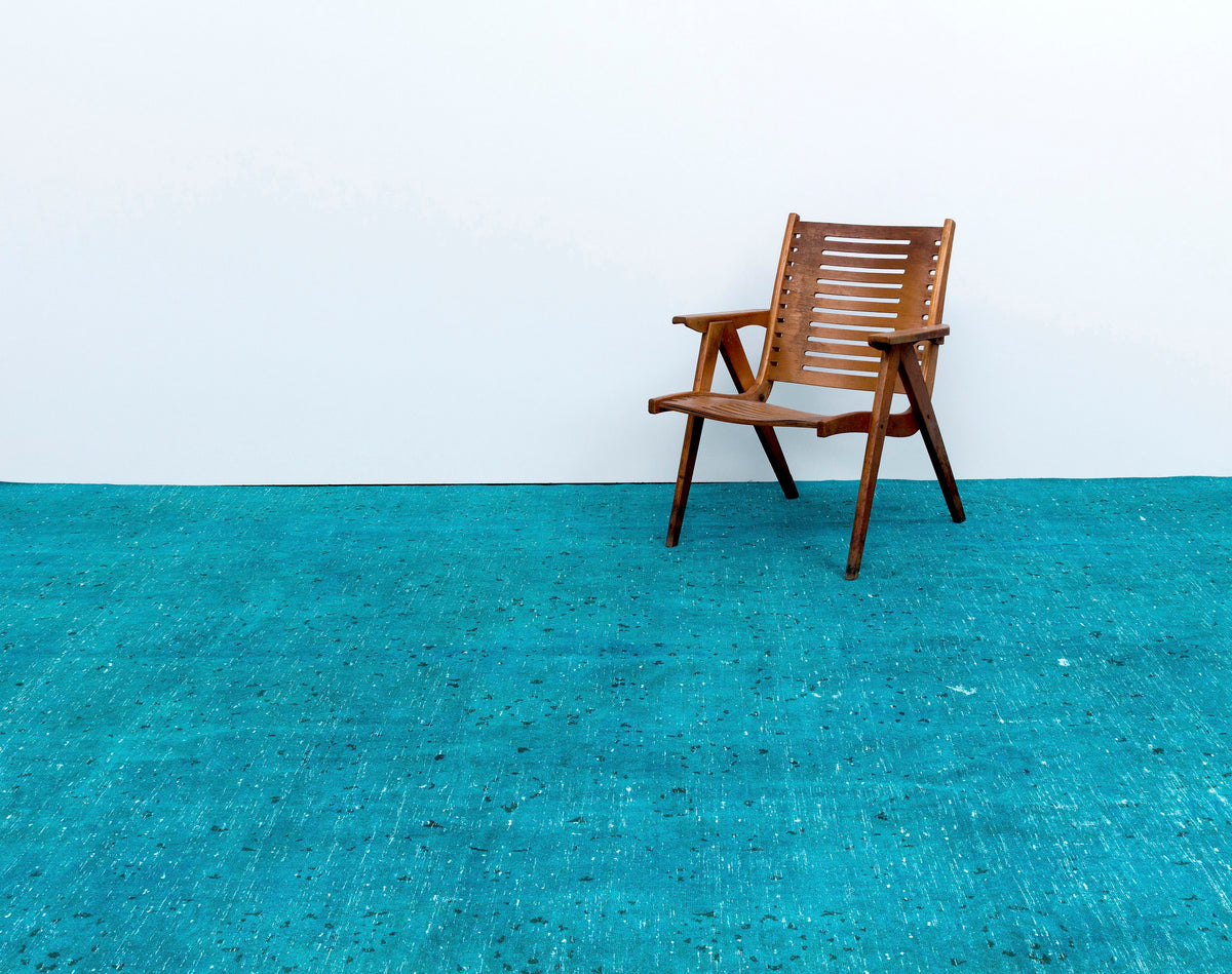 Solid turquoise rug with a distressed, speckled texture, styled with a wooden slatted chair against a white wall.