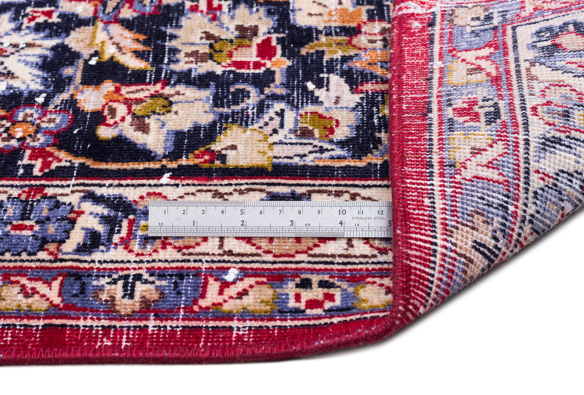 Traditional Persian-style rug with a navy, red, and cream floral pattern, shown folded with a metal ruler for scale.