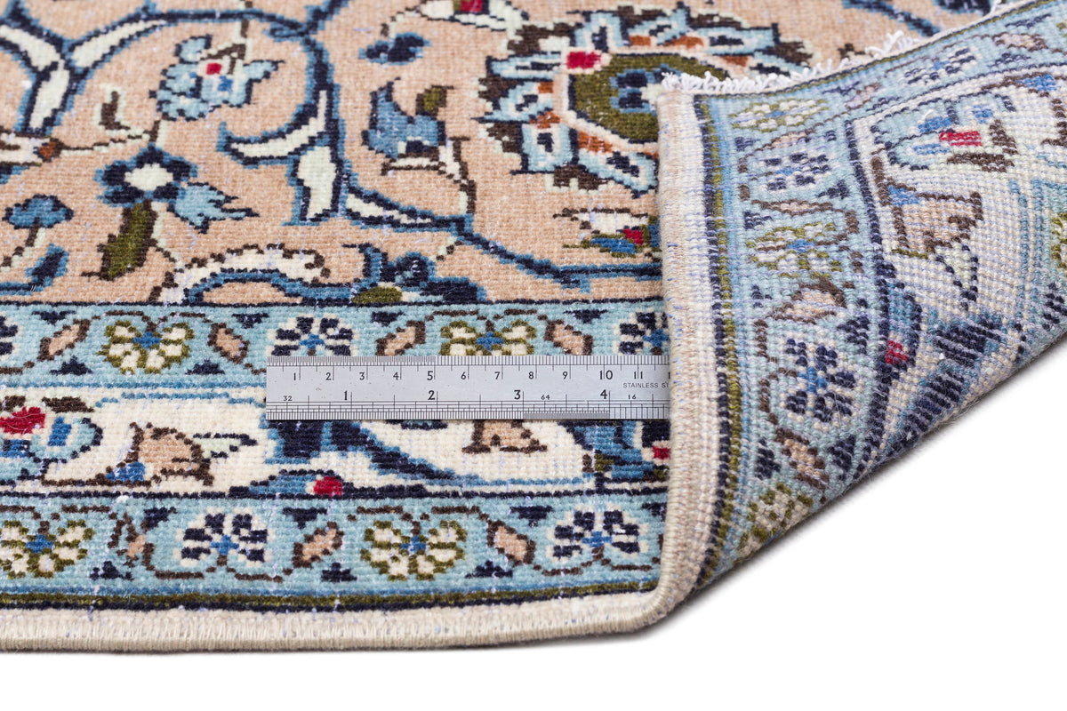 Beige and blue traditional floral rug with a folded corner and a metal ruler for scale on a white background.