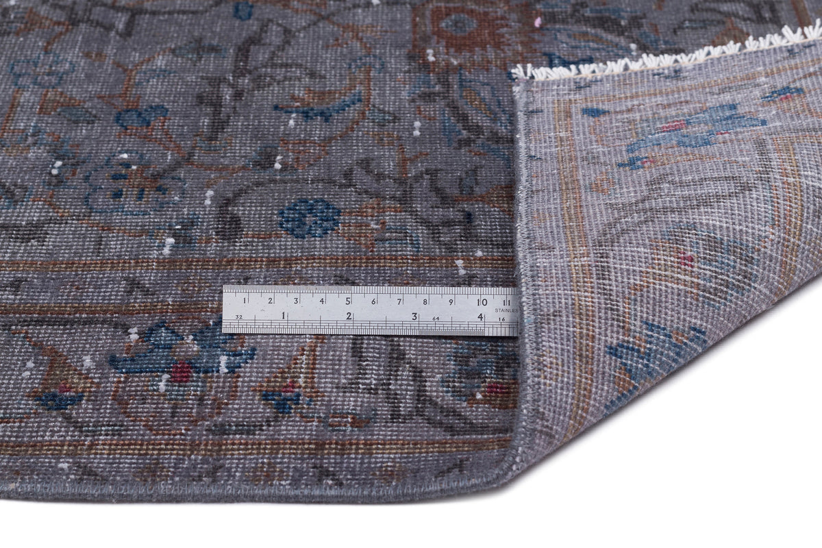 Grey traditional rug with blue and rust floral patterns, shown folded with a metal ruler for scale on a white background.