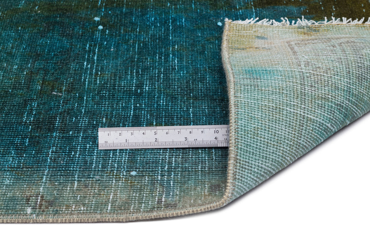 Teal and olive green distressed rug with a folded corner and a metal ruler for scale on a white background.