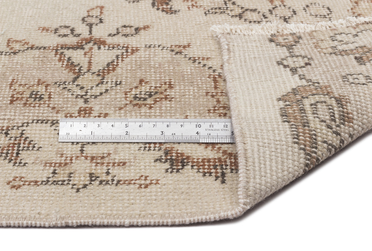 Beige rug with a brown and grey distressed floral pattern, shown with a metal ruler for scale on a white background.