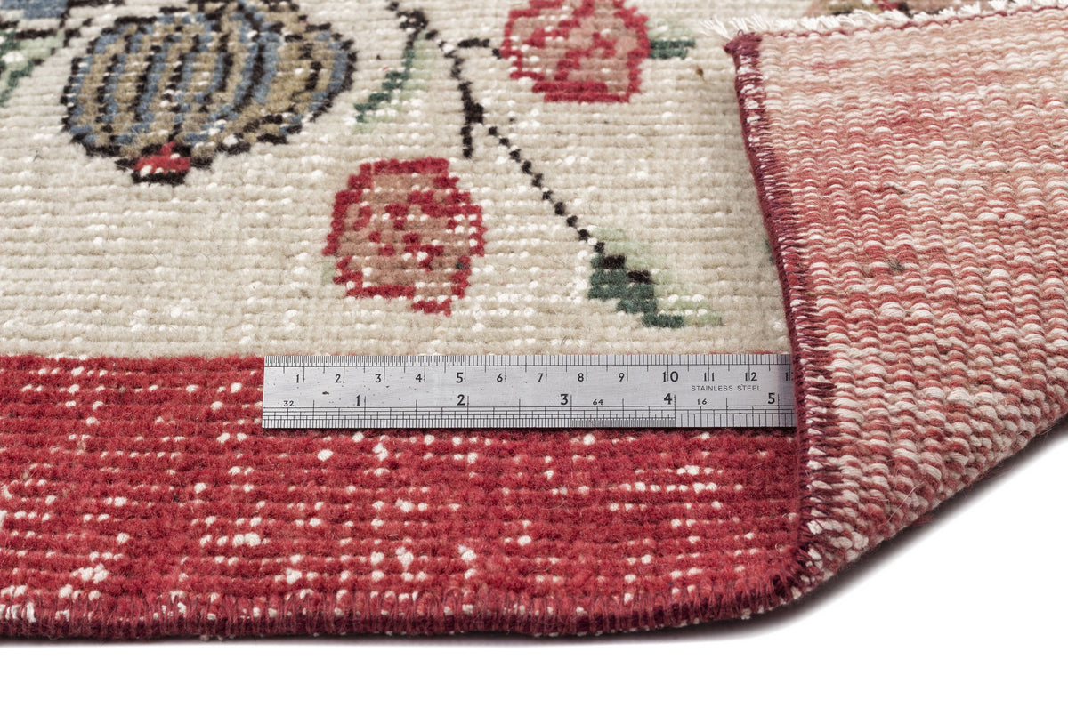 Cream rug with floral pattern, red border, and folded corner showing the underside, measured by a stainless steel ruler.