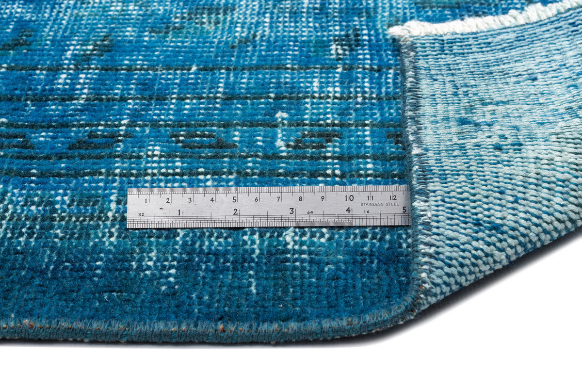 Distressed blue rug with a geometric grid pattern and a folded corner, shown with a metal ruler for scale.