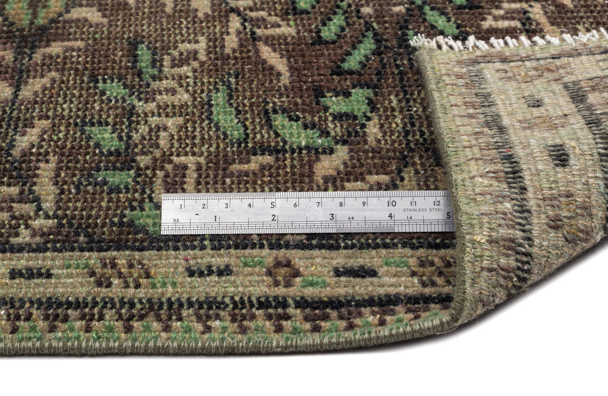 Brown and green traditional floral rug with a folded corner and a metal ruler for scale on a white background.