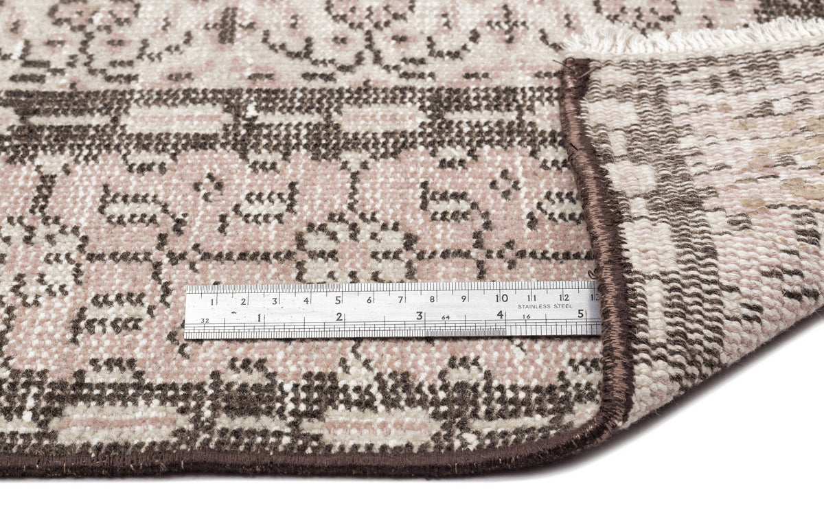 Pink and brown traditional patterned rug with a folded corner and a metal ruler for scale on a white background.