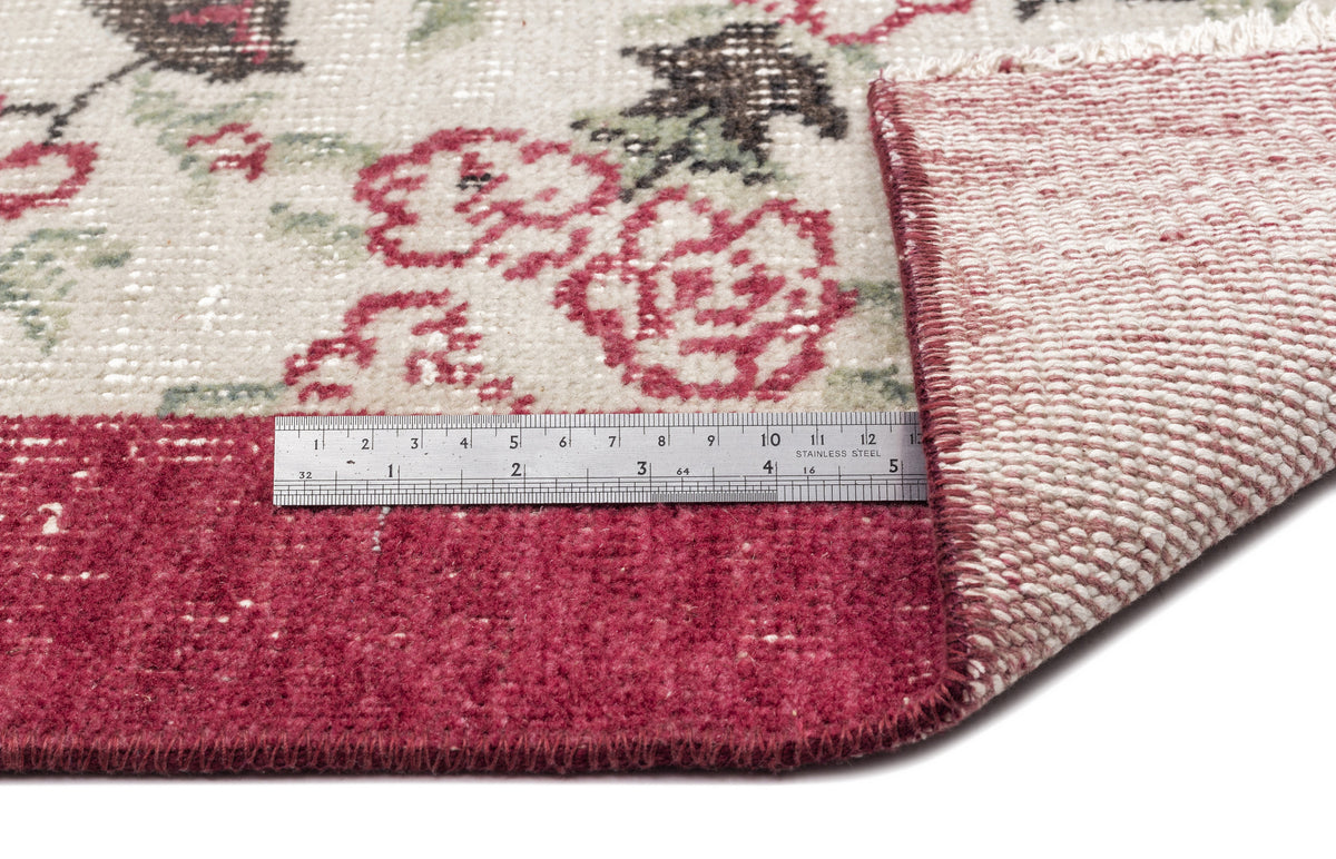 Cream rug with red floral pattern and solid red border, showing the woven backing and a metal ruler for scale.