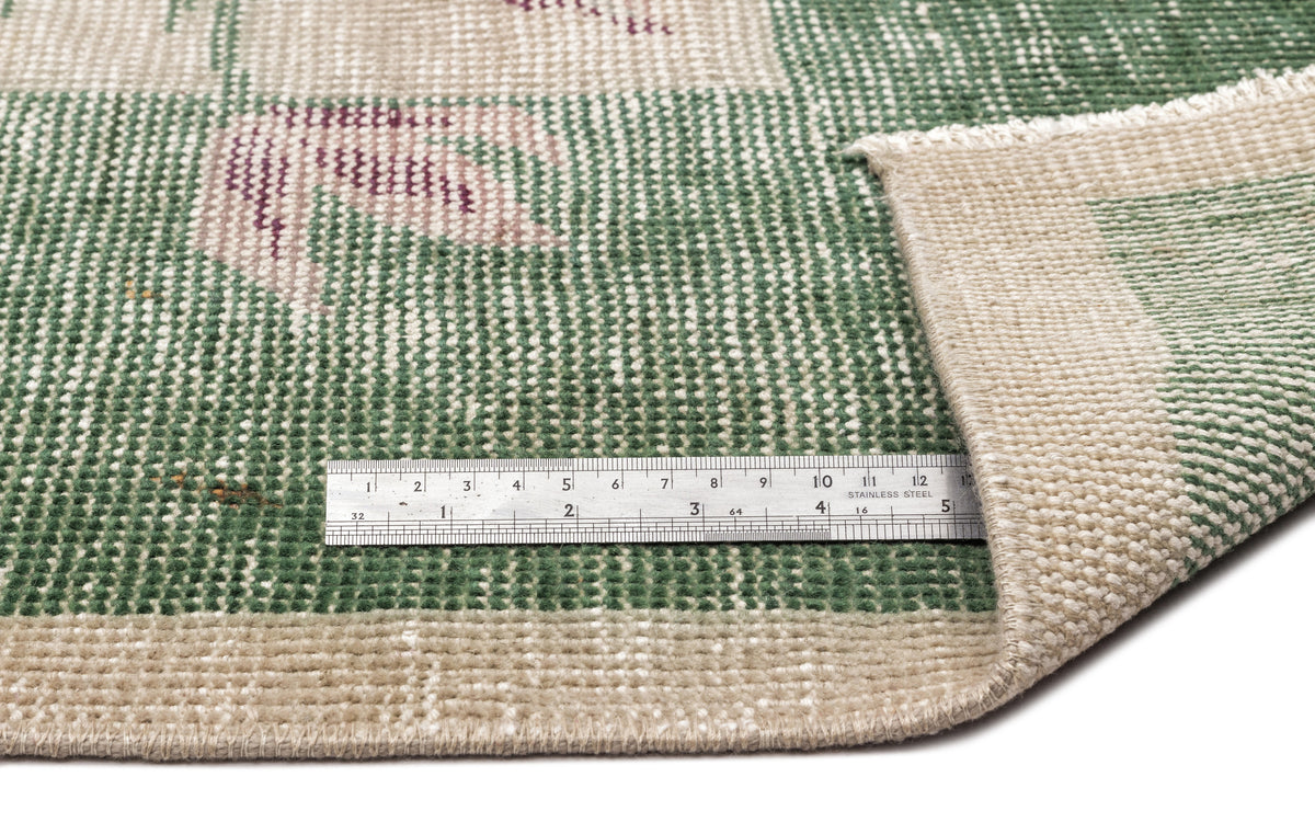 Green and beige woven rug with a floral pattern and a metal ruler for scale, shown on a white background.