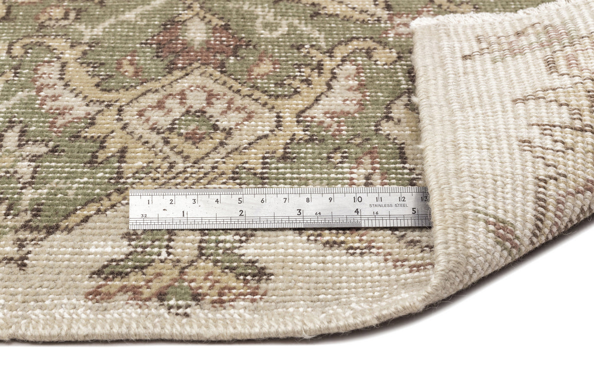 A close-up of the Seudian - Vintage Medallion Rug (5'9 X 8'9) shows its green, beige, and brown intricate pattern. A corner is flipped to reveal the back, and a metal ruler measuring about 6 inches provides scale.
