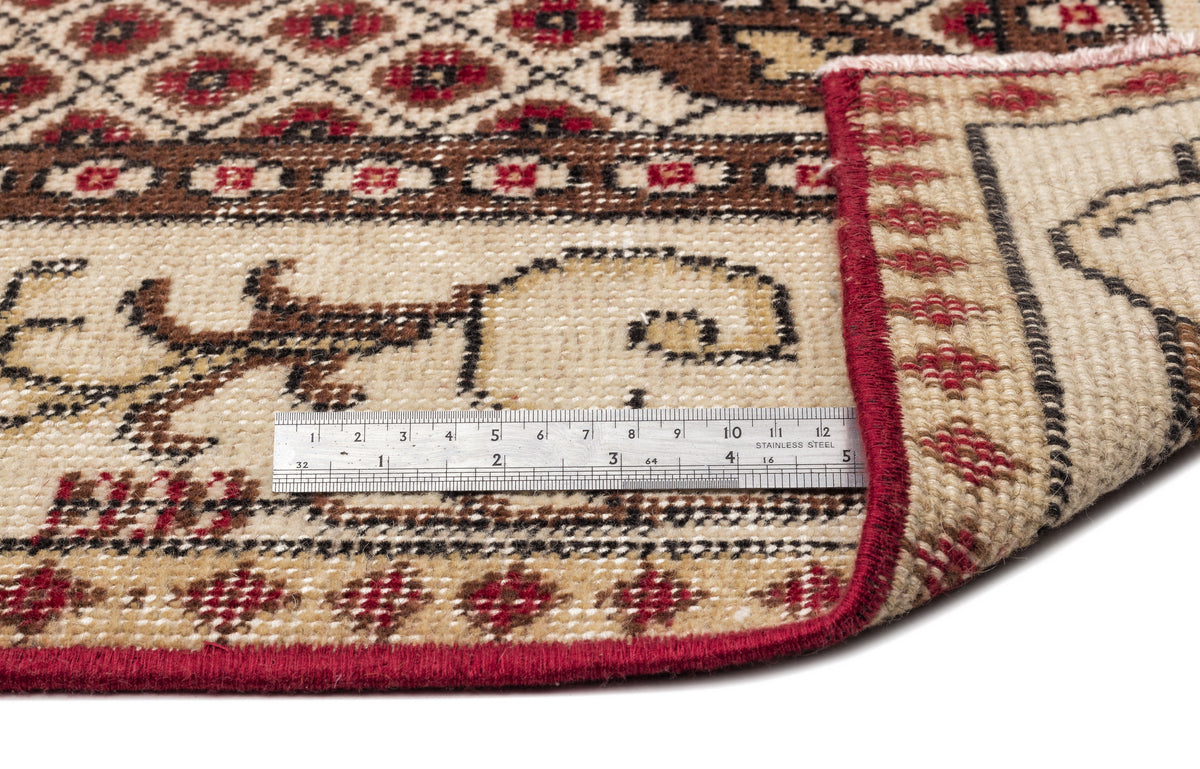 Beige, brown, and red traditional patterned rug with a folded corner and a metal ruler for scale on a white background.