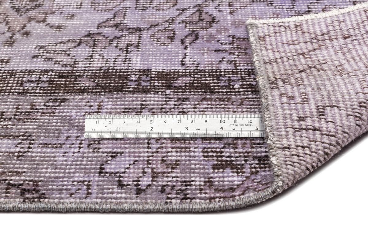 Lavender and brown distressed floral rug with a folded corner and a metal ruler for scale on a white background.