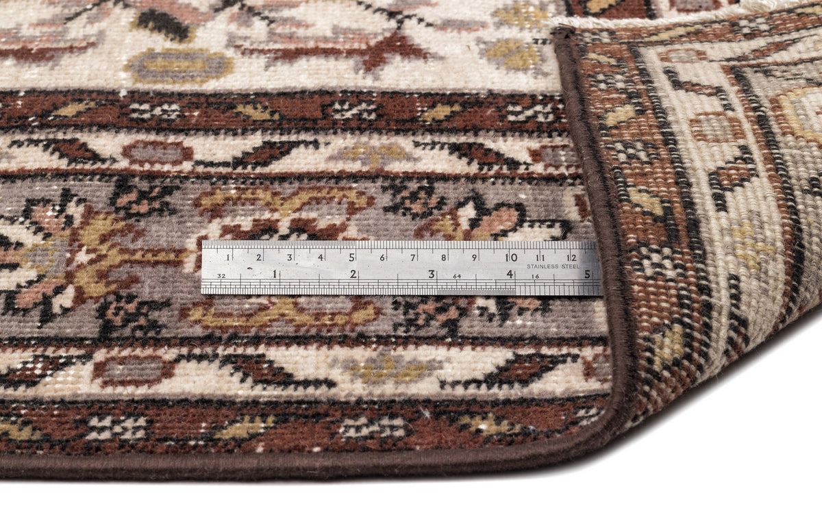 Traditional brown, cream, and grey patterned rug with a folded corner and a metal ruler for scale on a white background.