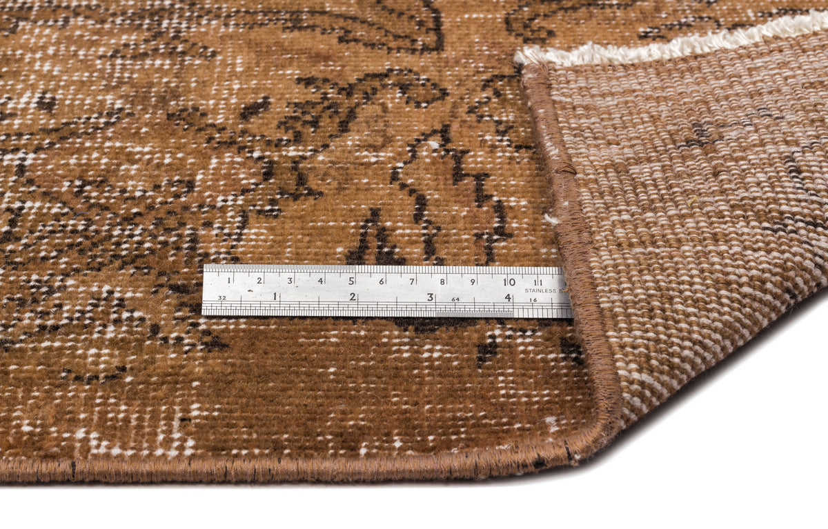 Brown and black distressed floral rug with a white fringe, shown with a metal ruler for scale on a white background.