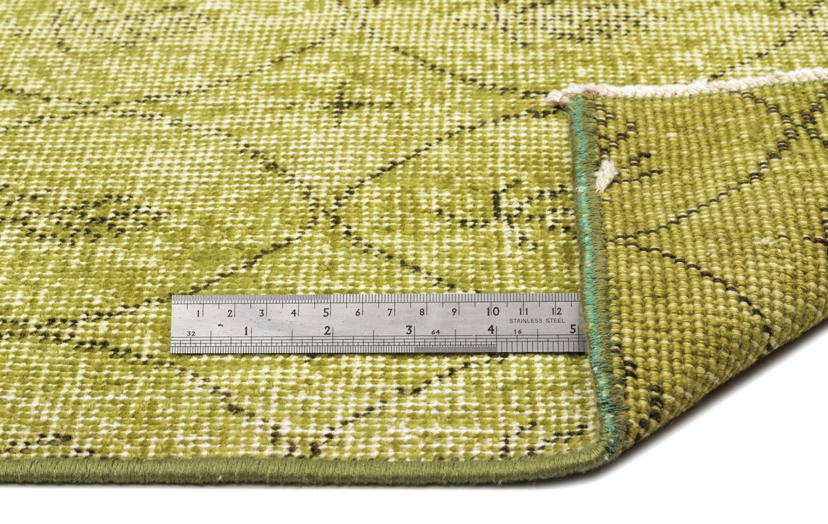 Olive green rug with a distressed diamond pattern and a metal ruler for scale, showing the texture and edge finish.