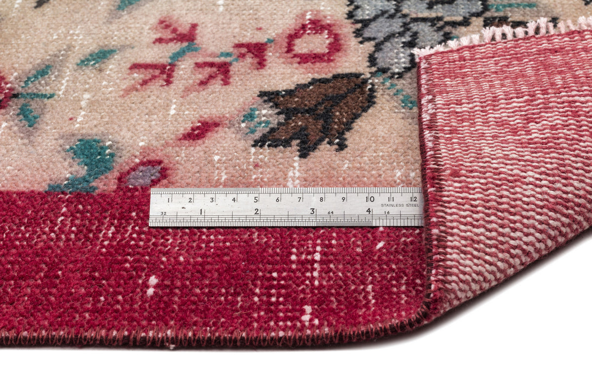 Beige and deep red floral rug with a distressed finish, showing the underside and a metal ruler for scale.
