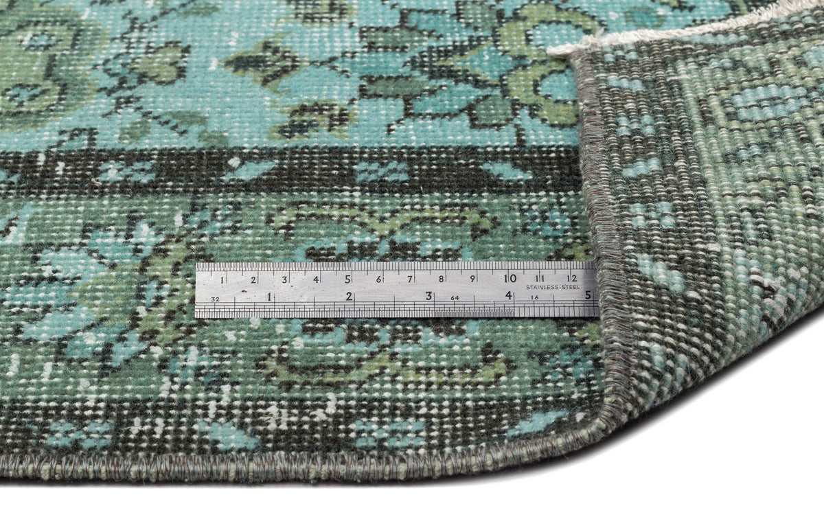 Teal and charcoal distressed floral rug with a folded corner and a metal ruler for scale on a white background.