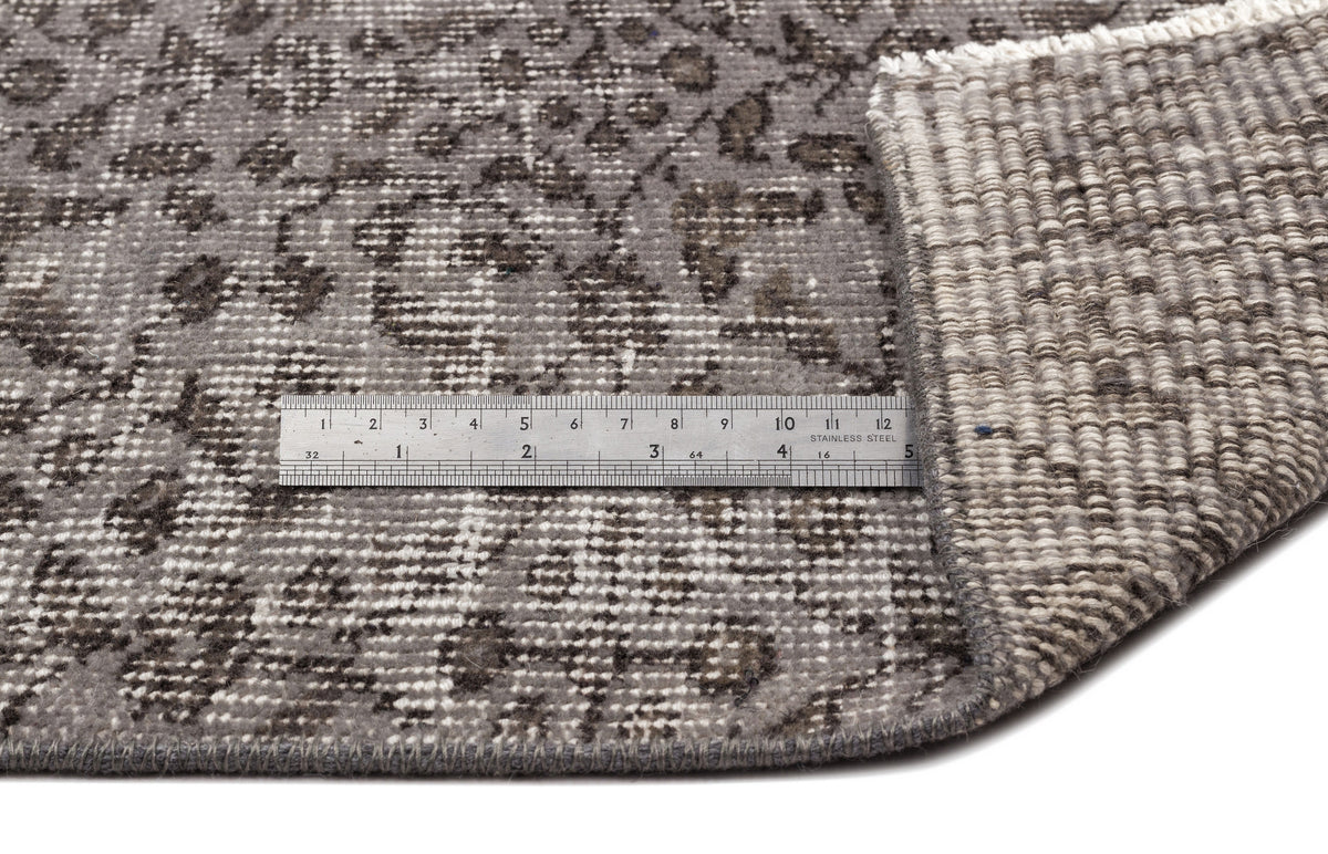 Grey and charcoal distressed floral pattern rug with a metal ruler showing pile height and backing detail.