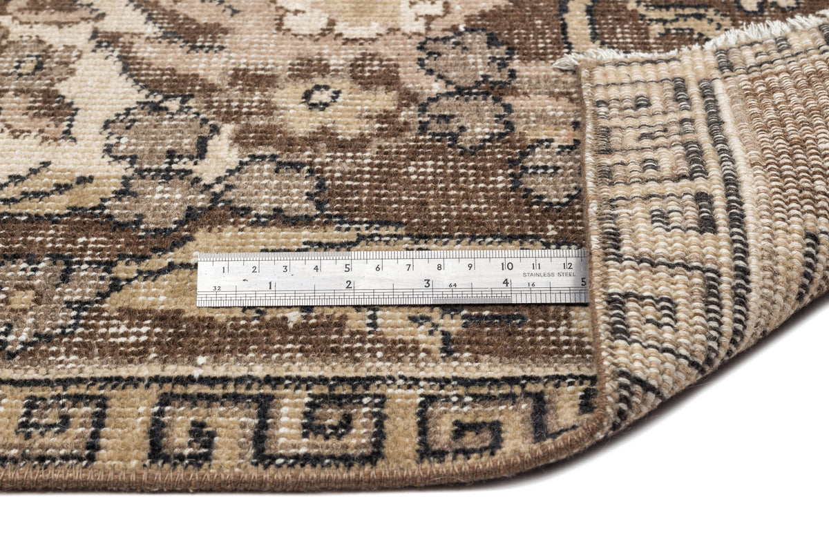 Brown and beige floral medallion rug with a folded corner and a stainless steel ruler for scale on a white background.