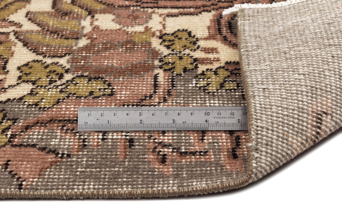 Vintage-style rug with a floral pattern in brown, olive, and cream, showing the woven backing and a metal ruler.