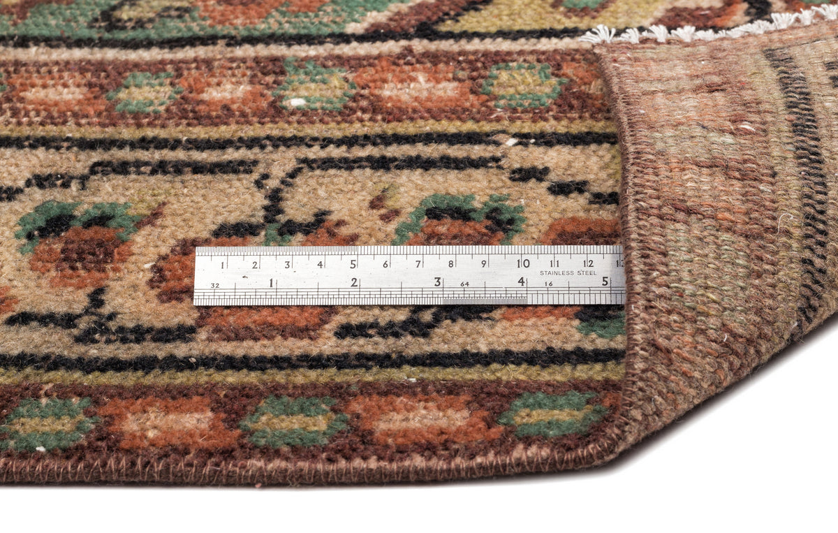 Close-up of a brown, tan, and green patterned rug with a metal ruler showing pile height, isolated on white.