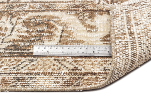 Load image 1 in gallery view. A metal ruler rests on the Tribuva - Vintage Medallion Rug - 6'3 x 9'6, highlighting its beige and brown pattern, thickness, and texture. One corner is folded to show the edge and underside of this vintage rug.