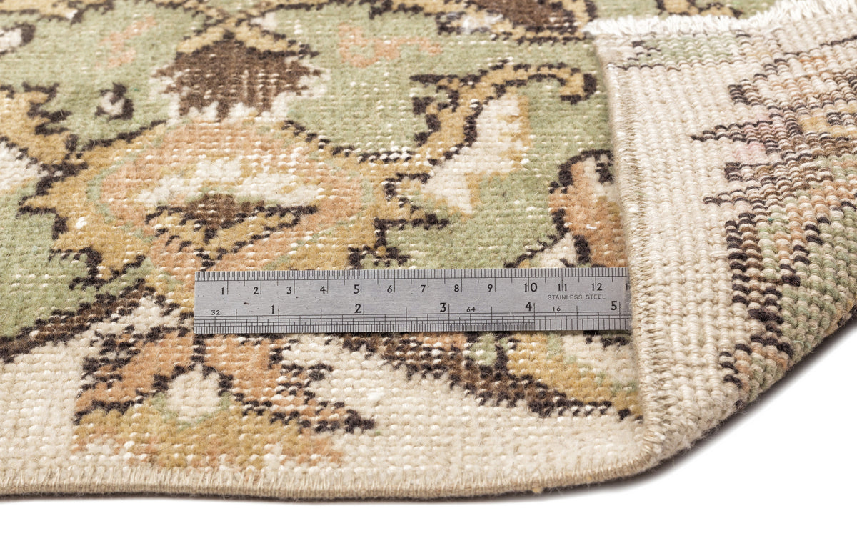 Sage green, tan, and brown floral patterned rug with a folded corner showing the woven backing and a metal ruler.