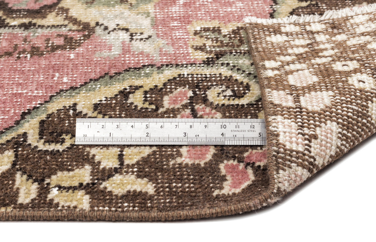 Distressed floral medallion rug in pink, brown, and cream with a metal ruler showing pile height on a white background.