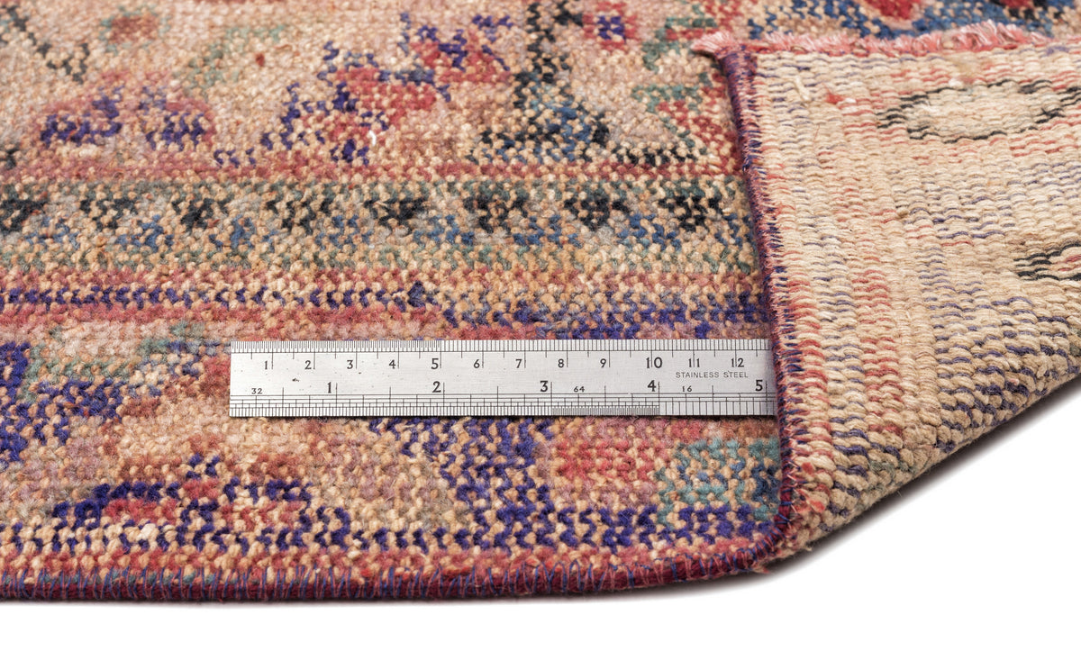 Distressed multi-colored rug with a floral pattern, shown folded to reveal the backing with a metal ruler for scale.