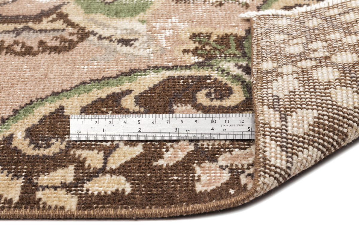 Distressed floral rug in brown, beige, and green with a metal ruler showing pile height and backing detail.